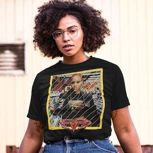 May include: Black t-shirt with a graphic of a woman with red hair, framed in yellow, and the text "POWER RECORDS". The shirt also displays a stylized logo in red and yellow. The model is wearing round glasses.