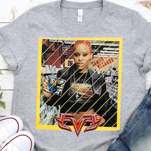 Op de afbeelding: Een grijs t-shirt met een afbeelding van de rapper Eve die een cd vasthoudt. De afbeelding staat in een gele lijst met diagonale lijnen. Het woord "EVE" wordt in een gestileerd, rood en zilver lettertype onderaan weergegeven.