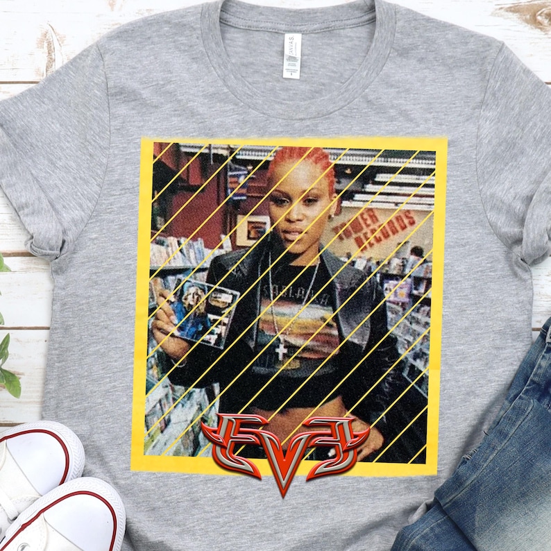 Op de afbeelding: Een grijs t-shirt met een afbeelding van de rapper Eve die een cd vasthoudt. De afbeelding staat in een gele lijst met diagonale lijnen. Het woord "EVE" wordt in een gestileerd, rood en zilver lettertype onderaan weergegeven.