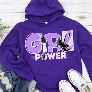 May include: A purple hooded sweatshirt with the words "GIRL POWER" in white and light purple. The letters "GIRL" are in a large, stylized font, with three superhero figures. The sweatshirt has a front pocket and a drawstring hood.