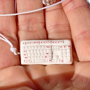 May include: A white miniature keyboard necklace with red keys. The necklace is on a white cord and is being held in a hand.