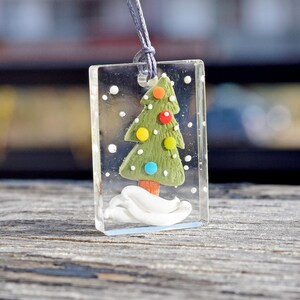 Puede incluir: Un colgante rectangular transparente con un árbol de Navidad verde en su interior. El árbol está decorado con adornos coloridos y se encuentra sobre una base blanca. El colgante está sobre una superficie de madera.