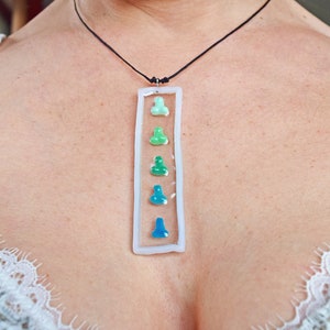 Könnte beinhalten: Eine weiße rechteckige Anhänger-Halskette mit einer Reihe von sechs kleinen, bunten Glasfiguren. Die Figuren sind in Grün- und Blautönen gehalten und erzeugen einen Farbverlauf.