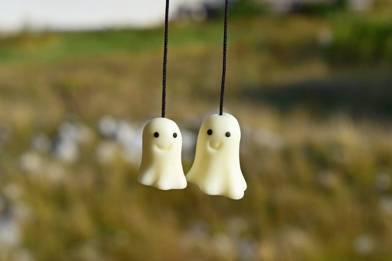 May include: Two white ghost-shaped charms with black eyes and smiling mouths hanging from black strings against a blurred green and brown background.