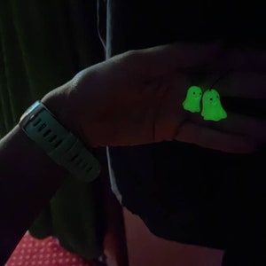 May include: A pair of glow-in-the-dark green ghost earrings. The earrings are shaped like ghosts with smiling faces.