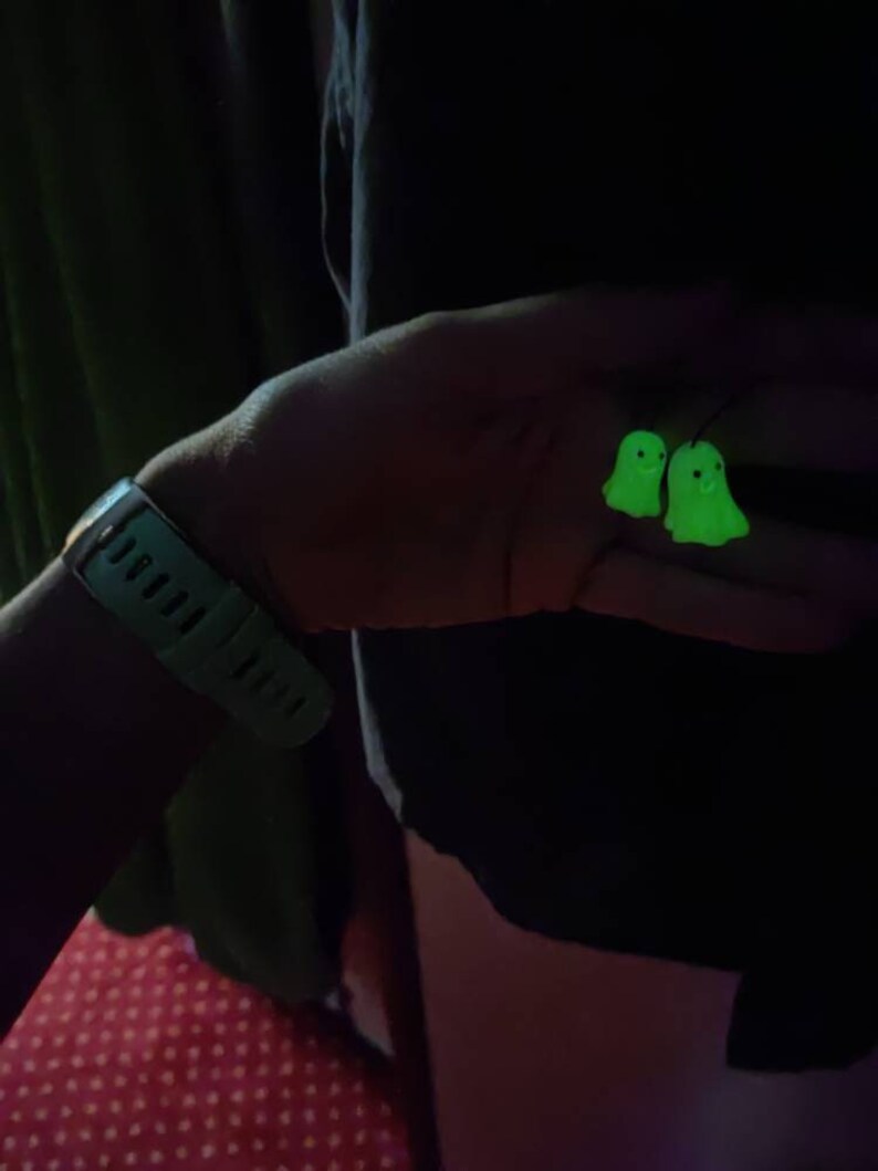 May include: A pair of glow-in-the-dark green ghost earrings. The earrings are shaped like ghosts with smiling faces.