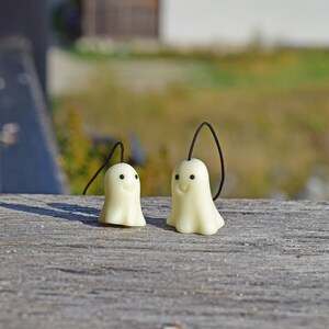 May include: Two white ghost-shaped charms with black eyes and smiling mouths hanging from black strings. The charms are on a wooden surface.