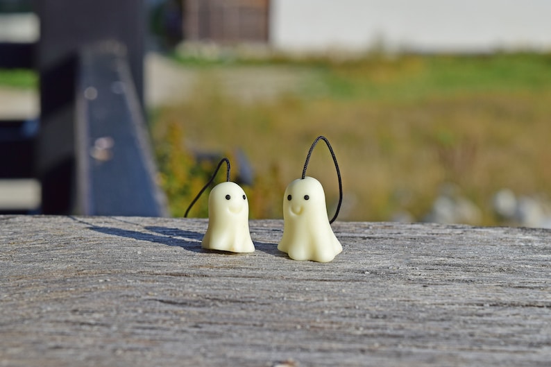 May include: Two white ghost-shaped charms with black eyes and smiling mouths hanging from black strings. The charms are on a wooden surface.
