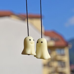 May include: Two white ghost-shaped ornaments with black eyes and smiling mouths hang from black strings against a blurred background of a building and blue sky.