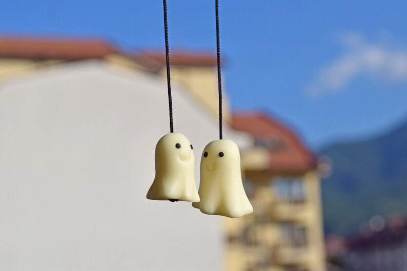May include: Two white ghost-shaped ornaments with black eyes and smiling mouths hang from black strings against a blurred background of a building and blue sky.
