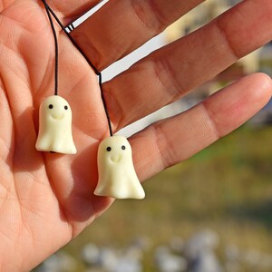 May include: Two small white ghost-shaped charms with black eyes and smiling mouths hanging from black strings. The charms are held in a person's hand.