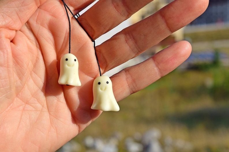 May include: Two small white ghost-shaped charms with black eyes and smiling mouths hanging from black strings. The charms are held in a person's hand.