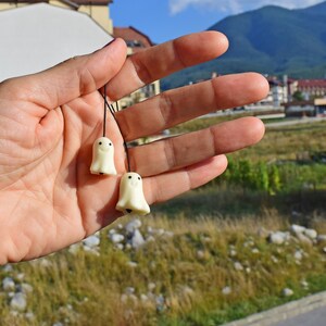 May include: Two small white ghost-shaped charms with black eyes and smiling mouths hanging from black strings. The charms are held in a person's hand.