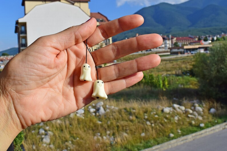 May include: Two small white ghost-shaped charms with black eyes and smiling mouths hanging from black strings. The charms are held in a person's hand.