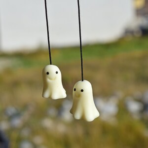 May include: Two white ghost-shaped ornaments with black eyes and smiling mouths hang from black strings against a blurred green background.