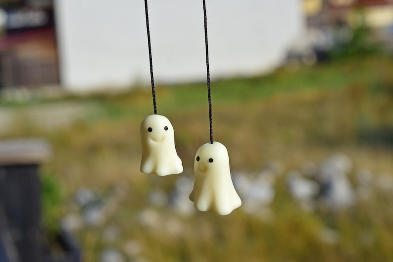 May include: Two white ghost-shaped ornaments with black eyes and smiling mouths hang from black strings against a blurred green background.