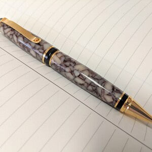 Könnte beinhalten: Ein weißer und brauner Marmor-Stift mit goldenen Akzenten und schwarzem Besatz. Der Stift hat eine goldene Klammer und eine goldene Spitze.