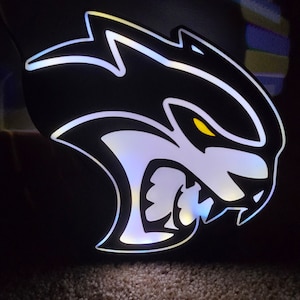 May include: A black and white illuminated sign with a snarling, stylized animal head. The animal's eyes are yellow and the sign is lit with white LEDs.