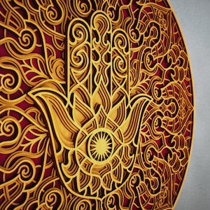 Könnte beinhalten: Eine runde Wanddekoration aus Holz mit einem detaillierten, komplizierten Design. Das Design zeigt eine große, stilisierte Hand mit einem Auge in der Mitte, umgeben von wirbelnden Mustern in Gold- und Rottönen.