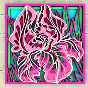 Peut inclure: Une découpe en papier rose et verte d'une fleur d'iris avec des détails complexes. La fleur est encadrée dans un carré rose avec un fond vert et bleu.