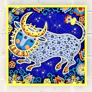 Op de afbeelding: Een blauwe stier met gele hoorns en een halve maan op zijn kop, omringd door kleurrijke bloemen en een sterrenhemel. De stier is een papieren uitsnijding met ingewikkelde details.