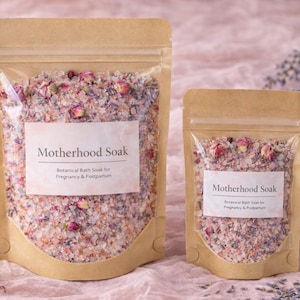 May include: Two kraft paper pouches filled with botanical bath soak. The larger pouch is filled with a mix of pink, white, and tan granules and dried rose buds. The smaller pouch has the same contents. Both pouches have a label that reads "Motherhood Soak".