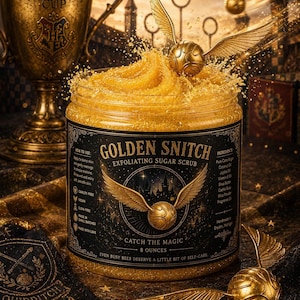 May include: A clear plastic jar of golden exfoliating sugar scrub labeled Golden Snitch, featuring a winged ball graphic and text including 8 ounces. The jar is surrounded by a golden trophy, a winged ball, and dark fabric with gold accents.