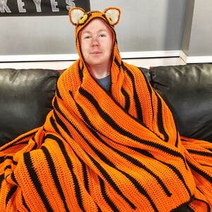 Peut inclure: Une personne portant une couverture en crochet ray&eacute;e orange et noire &agrave; rayures de tigre avec une capuche en t&ecirc;te de tigre. La couverture est drap&eacute; sur les &eacute;paules de la personne et elle est assise sur un canap&eacute;.