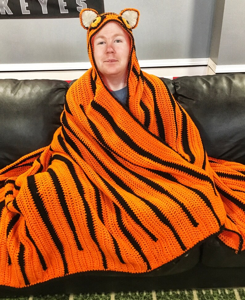 Peut inclure: Une personne portant une couverture en crochet ray&eacute;e orange et noire &agrave; rayures de tigre avec une capuche en t&ecirc;te de tigre. La couverture est drap&eacute; sur les &eacute;paules de la personne et elle est assise sur un canap&eacute;.