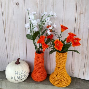 Könnte beinhalten: Zwei gehäkelte Vasen, eine orange und eine gelbe, mit weißen und orangefarbenen Blumen. Die Vasen stehen auf einer Holzoberfläche mit einem weißen Kürbis, auf dem "Hello Pumpkin" steht.
