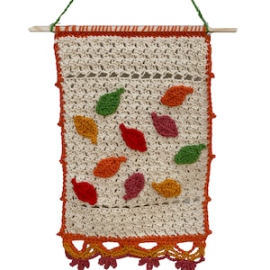 May include: A crocheted wall hanging with a cream background and colourful leaves in shades of orange, red, green, and pink. The hanging has a crocheted border in orange and a fringe at the bottom.