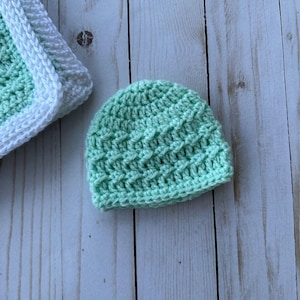 May include: A mint green crochet baby hat with a textured pattern. The hat is made of soft yarn and is likely handmade. A portion of a matching blanket with a white border is visible in the background.