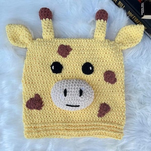 Op de afbeelding: Een handgemaakte gele gehaakte hoed in de vorm van een giraffekop. De hoed heeft bruine vlekken, zwarte ogen, een beige snuit en twee oren en hoorns met bruine uiteinden. De hoed is ontworpen om op het hoofd te worden gedragen.