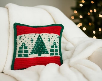 May include: A crocheted Christmas pillow with a red border and a green trim. The pillow features a white center with a green Christmas tree and gift design. The pillow is placed on a white blanket.