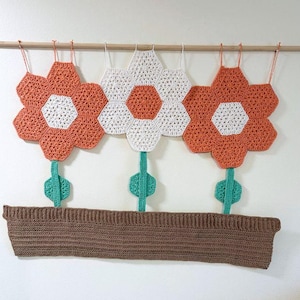 Peut inclure: Une tenture murale crochetée à la main représentant trois grandes fleurs orange et blanches, avec des tiges et des feuilles vertes. Les fleurs sont fixées à une jardinière marron et suspendues à une cheville en bois avec du fil orange et blanc.