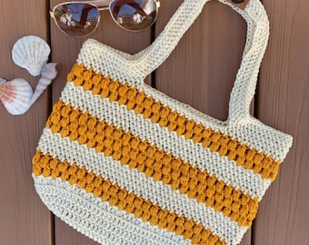 May include: A crocheted tote bag with white and mustard yellow stripes. The bag has a  crochet stitch that creates a bumpy texture. The bag has two straps and a brown leather tag with the letter "S" on it.
