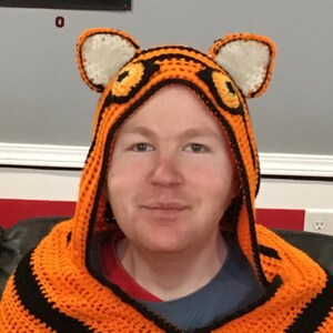 Peut inclure: Une personne portant une couverture &agrave; capuche en crochet &agrave; rayures orange et noires avec des oreilles de tigre.