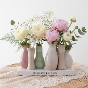 Könnte beinhalten: Ein Blumenarrangement mit verschiedenen Blumen in kleinen, pastellfarbenen Vasen. Das Arrangement umfasst rosa Pfingstrosen, weiße Rosen und Schleierkraut. Die Vasen sind auf einer weißen rechteckigen Basis mit dem Text "CELINA & TONI 06.10.2026" angeordnet.