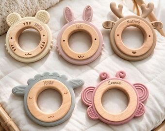 May include: Five circular baby teethers featuring wooden centers and silicone animal-shaped frames. The teethers are engraved with the names Adrian, Mila, Matheo, Jonah, and Leonie. Each piece displays distinct animal features like ears, antlers, or wings in cream, pink, brown, gray, and rose colors.