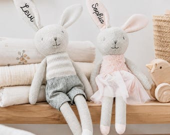 May include: Two knitted rabbit dolls with long ears. The left rabbit has the name Levi embroidered on its ear and wears grey shorts. The right rabbit has the name Sophia embroidered on its ear and wears a pink fuzzy top with a layered tulle skirt.