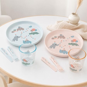 Può includere: Due set di stoviglie per bambini in azzurro e rosa tenue. Ogni set include un piatto rotondo, una forchetta, un coltello e un bicchiere trasparente. I piatti presentano illustrazioni di animali con cappellini da festa e festoni. I bicchieri sono personalizzati con i nomi Jonah e Leni in corsivo bianco.