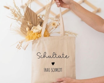 Könnte beinhalten: Eine beigefarbene Stofftasche mit dem schwarzen Aufdruck Schultüte über einem kleinen Herzsymbol und dem Namen FRAU SCHMIDT. Die Tasche ist mit einem dekorativen Arrangement aus getrockneten Gräsern, beigen Palmblättern und hellgelben Blumen gefüllt.