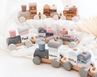 Op de afbeelding: Drie houten speelgoedtreinen in pastelkleuren, elk gepersonaliseerd met namen, geboortedata, tijden, gewichten en lengtes. De treinen bestaan uit stapelbare blokken met afgeronde randen, wielen en kleine pinnetjes, met details zoals Livia, Paul en Fabienne.