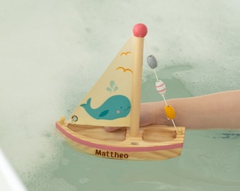 Può includere: Un giocattolo a forma di barca a vela in legno con un design a balena blu e una striscia rosa. La barca ha una corda di perline colorate attaccate all'albero maestro. La barca galleggia in una vasca da bagno con bolle. La barca è personalizzata con il nome "Mattheo".