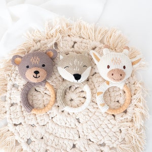 Può includere: Tre morsi di animali in maglia con anelli di legno. I morsi sono un orso marrone, una volpe grigia e una mucca bianca. I morsi sono su un tappetino in crochet beige.
