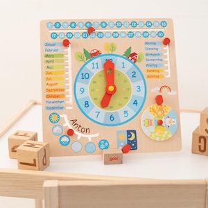 Könnte beinhalten: Lernuhr und Kalender aus Holz mit Zifferblatt, Monaten, Tagen und Wetterindikatoren. Die Uhr hat blaue Zahlen und rote Zeiger. Enthält Holzblöcke mit Buchstaben und dem Namen "Anton".