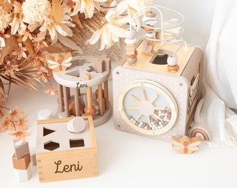 May include: Three wooden toys for babies and toddlers. The toys are in shades of brown and beige. The first toy is a shape sorter with the name "Leni" engraved on the front. The second toy is a spinning wheel with a sun design and beads. The third toy is a rotating cylinder with a butterfly design and the name "Leni" engraved on the side.