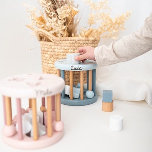 May include: Two wooden baby toys with spinning cylinders. The blue toy has the name "Luca" on it and the pink toy has the name "Mila" on it. The toys have different colored shapes inside the cylinders.