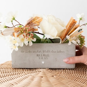 Könnte beinhalten: Ein rechteckiger Betonblumentopf gefüllt mit weißen Blumen, Grünzeug und gefaltetem Geld. Der Topf trägt die Inschrift "Wo Liebe wächst, blüht das Leben" und die Namen "MIRI & MIGUEL" mit einem Datum. Der Topf steht auf einer geflochtenen Korb-Oberfläche.