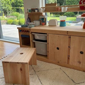 Peut inclure: Une cuisine jouet en bois avec un tabouret assorti. L'ensemble comprend une cuisinière, des armoires et des étagères avec des accessoires de jouets. Une fraise rouge et un champignon orange décorent le dessus. La cuisine est en bois clair.